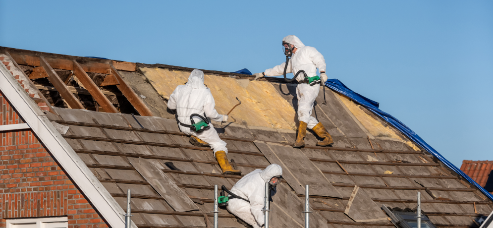 Three workers in protective suits remove shingles and insulation from a sloped roof under a clear blue sky.