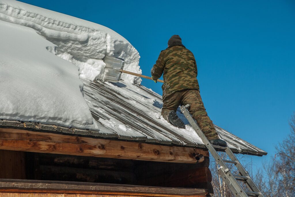 Worker in camouflage clothing clears heavy snow from the roof of a wooden structure, using a shovel on a ladder.