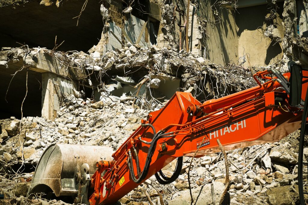 A demolition machine with an orange arm is working among rubble and debris from a partially destroyed building.