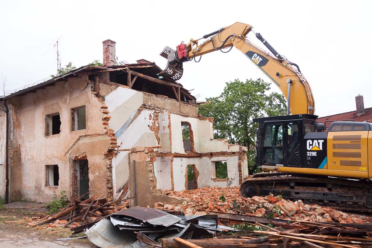 Yellow CAT excavator with grappling attachment demolishing a collapsed building, surrounded by debris and trees.