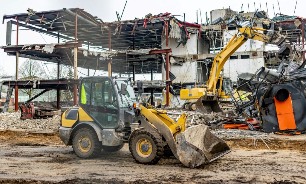 Excavators and a front loader at a demolition site, with debris and a partially collapsed building in the background.
