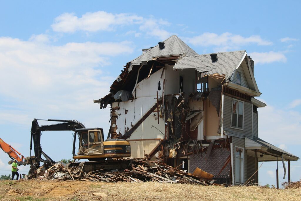Two-story house being demolished with exposed walls, debris on the ground, and a worker observing.